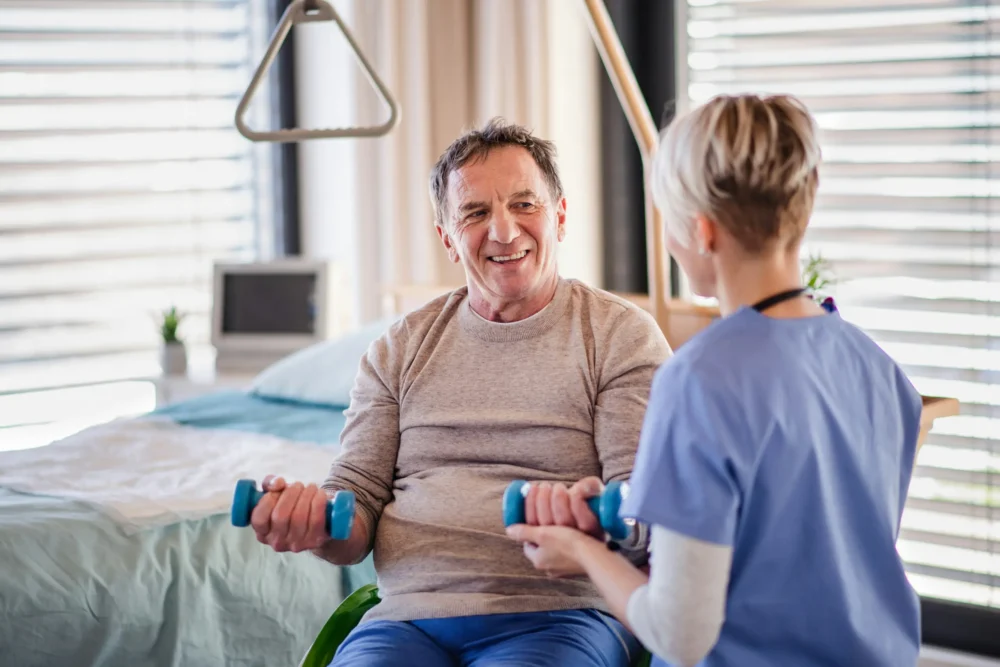 Older man lifting small weights in a hospital setting with a nurse helping. He is smiling.